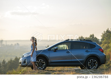 Happy woman driver in blue summer dress enjoying warm evening near her car. Travel and vacations concept. 136286788
