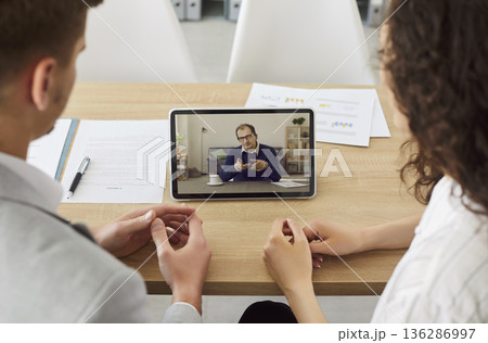 Company employees having online video call with colleague using tablet sitting in office. 136286997