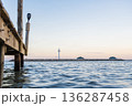Wooden Pier Over Calm Sea with Distant Lighthouse at Sunset 136287458