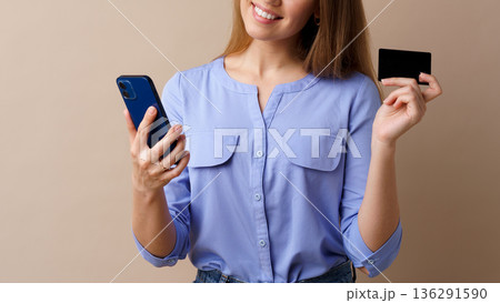 Woman holding a phone and a card while smiling in a simple indoor setting Woman holding a phone and a card while smiling in a simple indoor setting 136291590