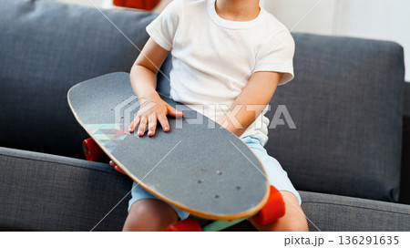 Child holds skateboard while sitting on sofa in a bright indoor space during the day 136291635