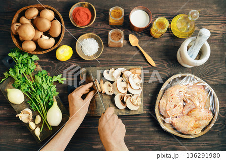 Top view of chef hands slicing fresh mushroom on wooden board with marinated chicken nearby 136291980