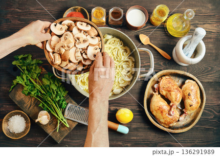 Hands adding sliced mushrooms to a pot with onions while cooking in a kitchen 136291989