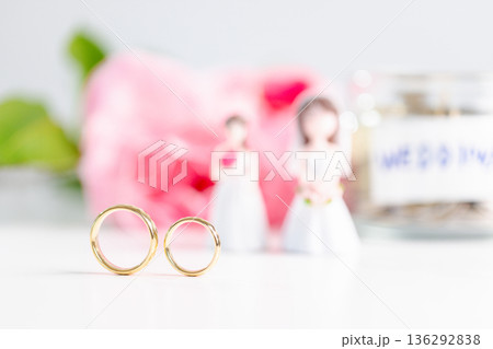 Inclusive wedding commitment concept celebrating equality, love and shared planning with pastel copy space; two gold rings in focus, blurred bride figurines, pink flowers and coin jar. celebrating Inclusive wedding commitment concept celebrating equality, love and shared planning with pastel copy space; two gold rings in focus, blurred bride figurines, pink flowers and coin jar. celebrating 136292838