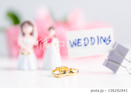 Inclusive wedding love concept celebrating diversity and equality with soft copy space for announcements; two gold rings in foreground, blurred bride figurines, WEDDING card and binder clips. Inclusive wedding love concept celebrating diversity and equality with soft copy space for announcements; two gold rings in foreground, blurred bride figurines, WEDDING card and binder clips. 136292841