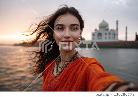 Woman in sari takes selfie with taj mahal at sunset, wearing traditional jewelry and vivid clothing 136296578