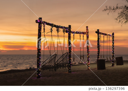 Swings are set up on the beach as the sun sets in the background. Lights hang from the swing posts, and the waves of the sea are visible nearby. High quality photo Swings are set up on the beach as the sun sets in the background. Lights hang from the swing posts, and the waves of the sea are visible nearby. High quality photo 136297396