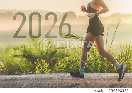Sports start and the start of 2026 new year. A woman jogs on a road at dawn while surrounded by green vegetation. The sun rises in the background, illuminating the scene and creating a fresh start to 136298380