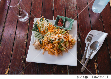 Pad Thai fried noodles on a white square plate on a wooden table in a rustic outdoor restaurant. Top view of Flat lay real dish in real life, authentic thailand. High quality photo 136298381