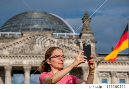 Berlin, germany, august 12, 2023. Woman tourist using a smartphone for capturing the reichstag building and fluttering german flag, experiencing travel in berlin 136300362