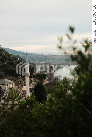 View of Pocitelj Fortress with mosque minaret and stone tower above the Neretva River valley in Bosnia and Herzegovina. Historic hillside settlement surrounded by mountains and winter vegetation 136300410