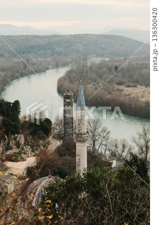 View of Pocitelj mosque minaret and historic stone tower above the Neretva River, framed by trees and hills in a peaceful cultural landscape 136300420