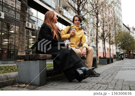 Two female friends sit on a bench with coffee cups, talking and enjoying the city surroundings. The concept highlights urban lifestyle, communication and companionship 136300636