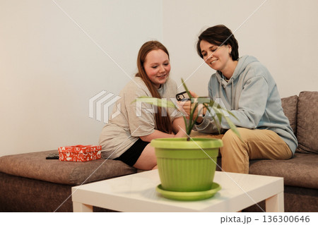 Two friends sit together on a couch and look at a smartphone, smiling at the screen. The concept represents digital connection, friendship and shared moments 136300646