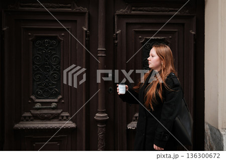 A young woman walks along the street holding a coffee cup near an ornate vintage door. The concept conveys everyday life, movement and an urban cultural atmosphere A young woman walks along the street holding a coffee cup near an ornate vintage door. The concept conveys everyday life, movement and an urban cultural atmosphere 136300672