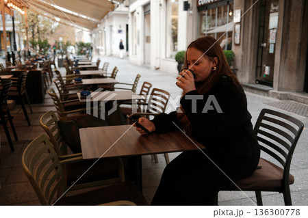 A young woman drinks coffee while sitting at an outdoor cafe table and holding her phone. The concept shows relaxation, slow living and everyday urban comfort 136300778