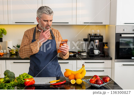 Middle-aged man shocked by bad news on smartphone in home kitchen after sudden internet message loss 136300827