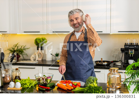 Middle-aged man chopping red pepper wearing headphones enjoying music while preparing healthy meal Middle-aged man chopping red pepper wearing headphones enjoying music while preparing healthy meal 136300831