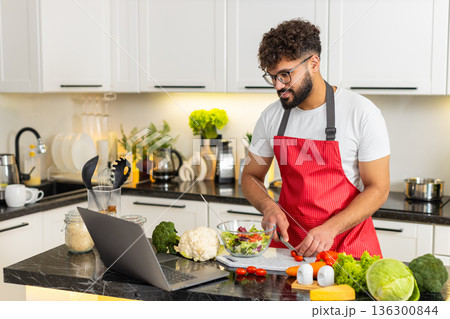 Arabian young adult man cooking fresh salad in kitchen watching recipe on laptop for quick meal prep 136300844