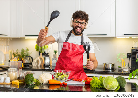 Arabian young adult man dancing in kitchen cooking salad listening to music in wireless headphones 136300848