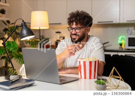 Arabian man plays movie trailer on laptop eats popcorn laughs aloud enjoys preview at night indoors 136300864
