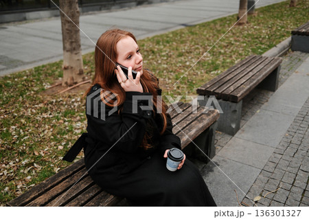 A woman in a black coat sits on a wooden bench outdoors, holding a coffee cup and talking on her smartphone during a chilly autumn day A woman in a black coat sits on a wooden bench outdoors, holding a coffee cup and talking on her smartphone during a chilly autumn day 136301327