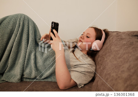 Girl lying on a couch with pink headphones, covered with a soft blanket, watching her phone. Cozy and peaceful indoor moment of evening relaxation Girl lying on a couch with pink headphones, covered with a soft blanket, watching her phone. Cozy and peaceful indoor moment of evening relaxation 136301368