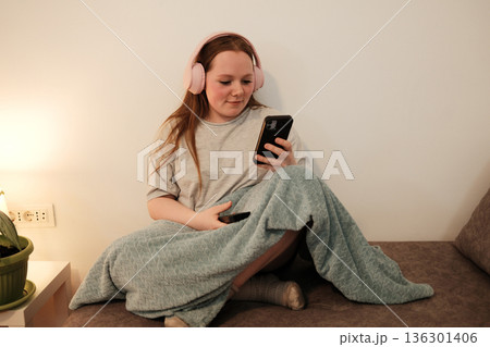 A woman with pink headphones sits on the sofa and looks at her smartphone, relaxing under a blanket in a cozy room 136301406