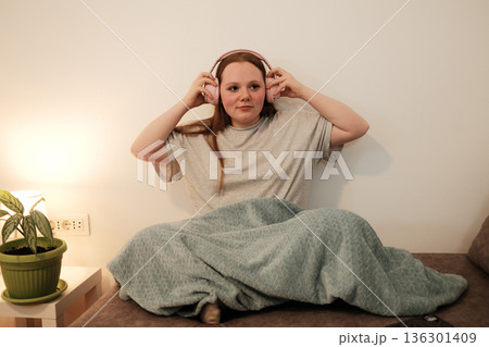 A woman sits on a sofa and puts on pink headphones, preparing to listen to music or audio while relaxing under a blanket A woman sits on a sofa and puts on pink headphones, preparing to listen to music or audio while relaxing under a blanket 136301409