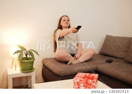 A woman sits cross-legged on a sofa and uses a remote control to switch channels, creating a cozy evening atmosphere at home A woman sits cross-legged on a sofa and uses a remote control to switch channels, creating a cozy evening atmosphere at home 136301413