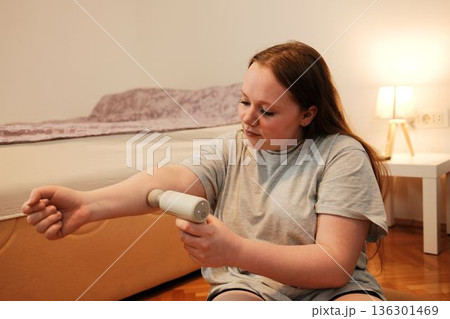 A woman applies a small handheld massage device to her arm, showing a simple recovery and self-care moment at home 136301469