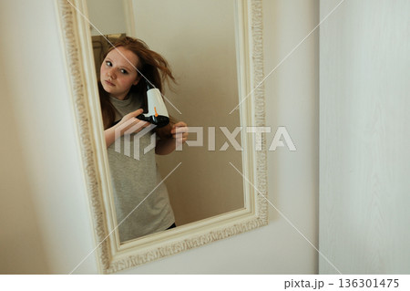 A woman uses a hairdryer while watching her reflection in the mirror, completing her morning grooming routine at home 136301475