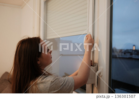 A woman stands by the window and raises the blind, letting in natural daylight and fresh air, symbolizing the beginning of a new day at home 136301507