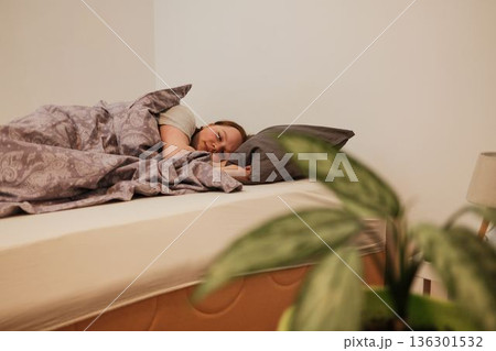A woman sleeps comfortably in a cozy bedroom, wrapped in a patterned blanket, creating a calm morning atmosphere and a sense of home comfort 136301532