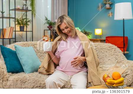Pregnant woman in blanket sipping herbal tea humming lullaby to baby enjoying cozy soft calm evening 136301579