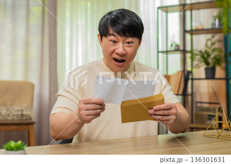 Asian man smiling while reading open letter on paper, glad about good news, joyful reaction at desk Asian man smiling while reading open letter on paper, glad about good news, joyful reaction at desk 136301631