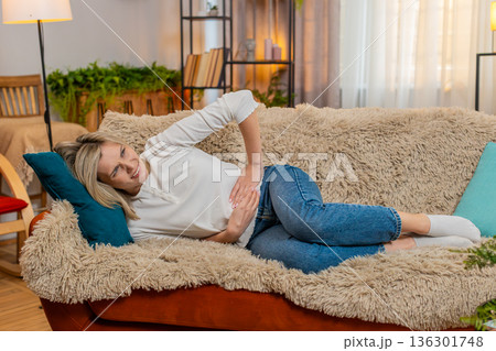 Adult woman holding stomach at home, expressing abdominal pain and discomfort while lying on sofa Adult woman holding stomach at home, expressing abdominal pain and discomfort while lying on sofa 136301748