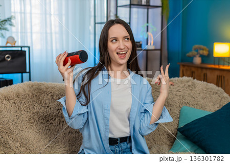 Happy woman dancing on sofa with wireless speaker laughing enjoying music raising hands rock gesture Happy woman dancing on sofa with wireless speaker laughing enjoying music raising hands rock gesture 136301782