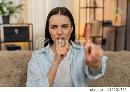 Young woman sitting on sofa making silence gesture placing finger to lips quietly saying shh secret 136301785