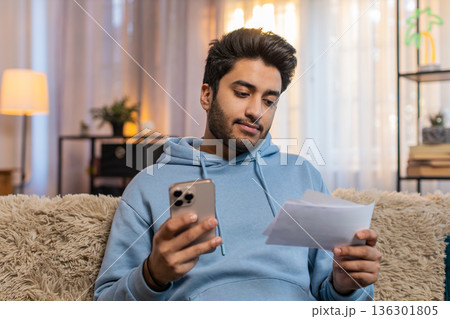 Young Indian man paying utility bills on smartphone with receipts sitting on sofa in living room Young Indian man paying utility bills on smartphone with receipts sitting on sofa in living room 136301805
