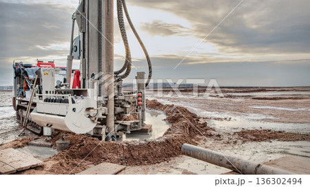 A drilling rig is busy extracting oil and gas resources in a vast, muddy area under an overcast sky. Surroundings show signs of drilling activity and equipment A drilling rig is busy extracting oil and gas resources in a vast, muddy area under an overcast sky. Surroundings show signs of drilling activity and equipment 136302494