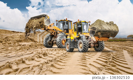 Two powerful loaders carry buckets of earth and sand to level the construction site, while a large pile of sand can be seen in the background, indicating ongoing construction work 136302510