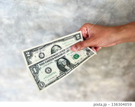 Hand holding a one US dollar banknote against a neutral concrete background, symbolizing cash payment, personal finance, small budget, inflation awareness, savings mindset, and everyday money value. Hand holding a one US dollar banknote against a neutral concrete background, symbolizing cash payment, personal finance, small budget, inflation awareness, savings mindset, and everyday money value. 136304059