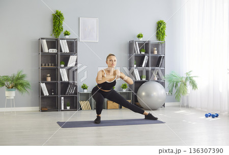 Woman performing side lunge home workout on yoga mat in bright home studio Woman performing side lunge home workout on yoga mat in bright home studio 136307390