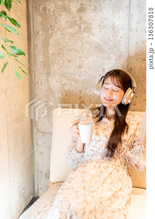ソファーに座りヘッドホンで音楽を聴きながらコーヒーを飲んでくつろいでいる若い女性 ソファーに座りヘッドホンで音楽を聴きながらコーヒーを飲んでくつろいでいる若い女性 136307818