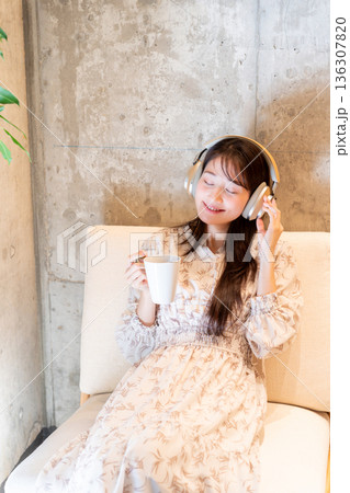 ソファーに座りヘッドホンで音楽を聴きながらコーヒーを飲んでくつろいでいる若い女性 ソファーに座りヘッドホンで音楽を聴きながらコーヒーを飲んでくつろいでいる若い女性 136307820