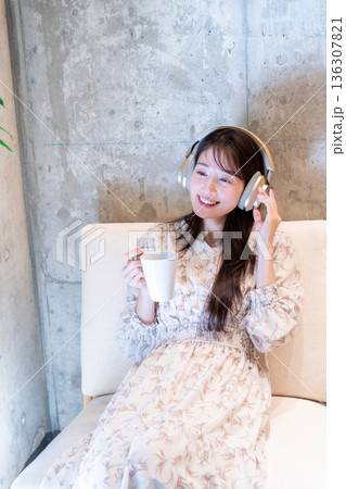 ソファーに座りヘッドホンで音楽を聴きながらコーヒーを飲んでくつろいでいる若い女性 ソファーに座りヘッドホンで音楽を聴きながらコーヒーを飲んでくつろいでいる若い女性 136307821