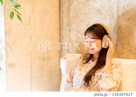 ソファーに座りヘッドホンで音楽を聴きながらコーヒーを飲んでくつろいでいる若い女性 ソファーに座りヘッドホンで音楽を聴きながらコーヒーを飲んでくつろいでいる若い女性 136307827