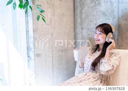 ソファーに座りヘッドホンで音楽を聴きながらコーヒーを飲んでくつろいでいる若い女性 136307828