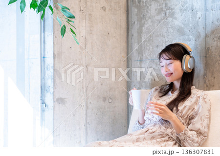 ソファーに座りヘッドホンで音楽を聴きながらコーヒーを飲んでくつろいでいる若い女性 ソファーに座りヘッドホンで音楽を聴きながらコーヒーを飲んでくつろいでいる若い女性 136307831